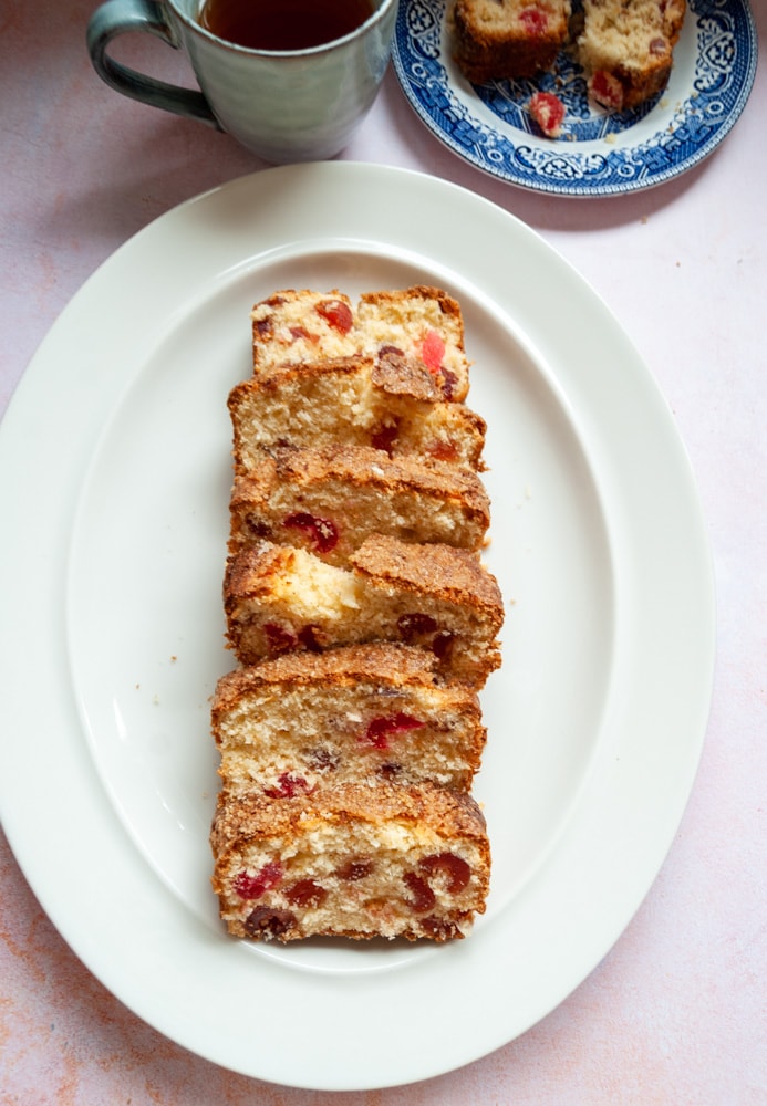 slices of cherry loaf cake on an oval white plate and pink backdrop. A blue stoneware mug of tea and a blue willow patterned plate with a slice of the cherry cake