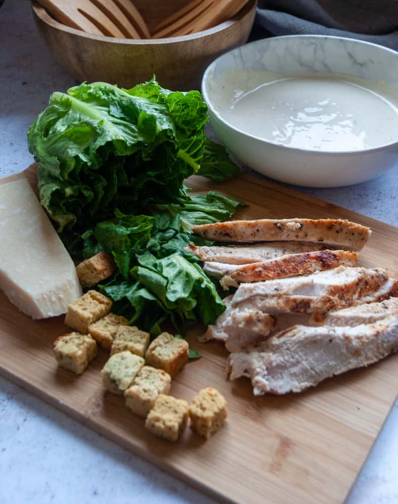 griddled chicken strips, a romaine lettuce, parmesan cheese, croutons and a white bowl of Caesar dressing on a wooden board.