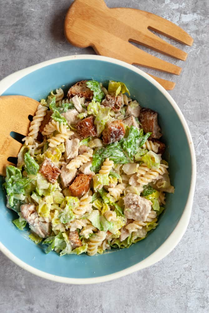 a blue and white bowl filled with chicken Caesar salad with croutons and pasta with wooden salad servers 