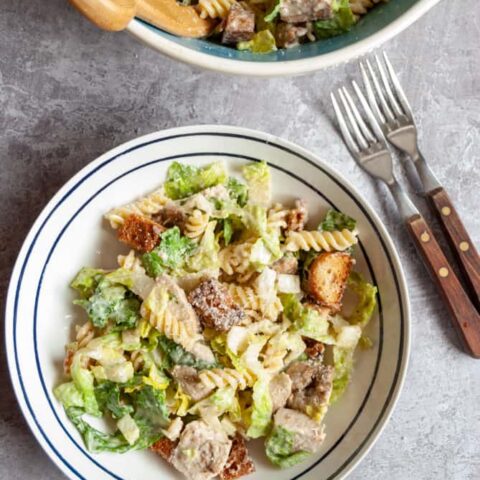 a white serving bowl of Caesar salad with chicken, croutons and pasta and a wooden handled knife and fork.