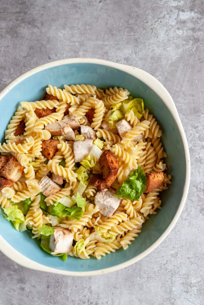 a blue and white bowl of Caesar salad with chicken, pasta and croutons on a grey backdrop.