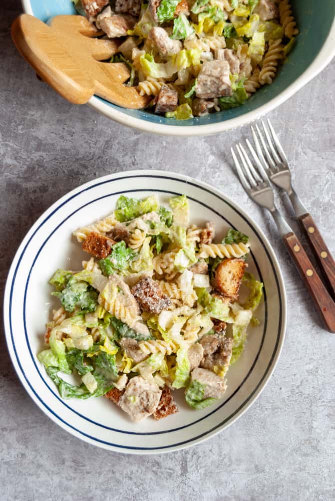 a white serving bowl of Caesar salad with chicken, croutons and pasta and a wooden handled knife and fork.  