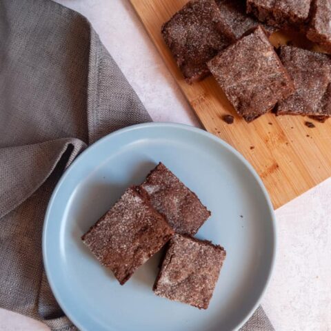 an overhead image of three chocolate slices sprinkled with sugar on a blue plate with a grey napkin, a blue mug of tea and a wooden board with more chocolate slices.