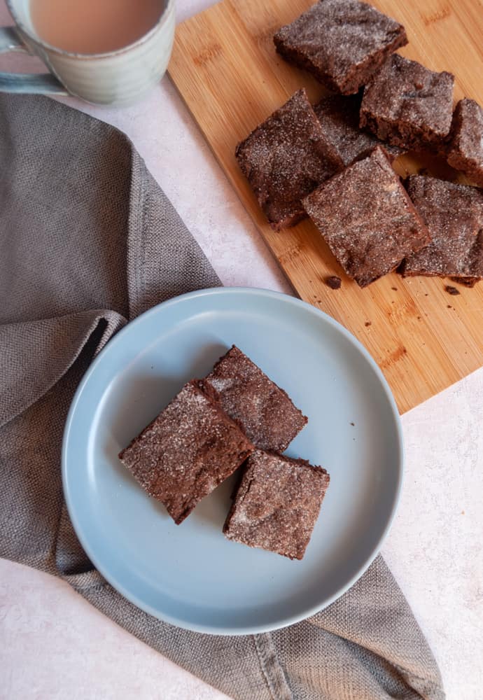 an overhead image of three chocolate slices sprinkled with sugar on a blue plate with a grey napkin, a blue mug of tea and a wooden board with more chocolate slices.