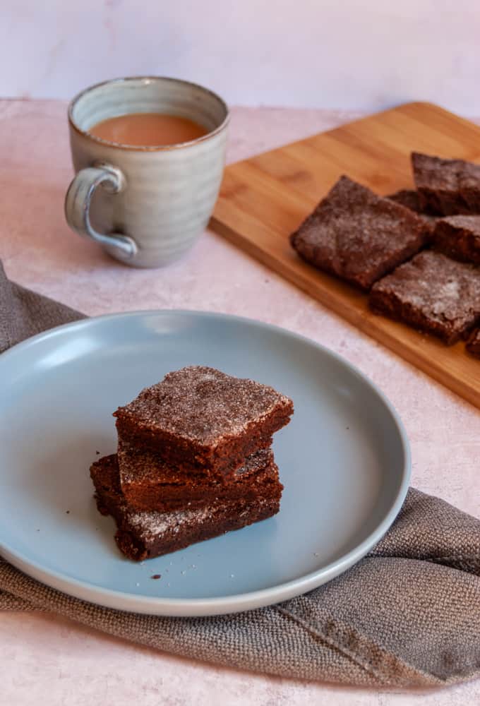 A blue plate with three chocolate slices sprinkled with sugar sitting on a grey napkin, a blue mug of tea and a wooden chopping board with more chocolate slices.