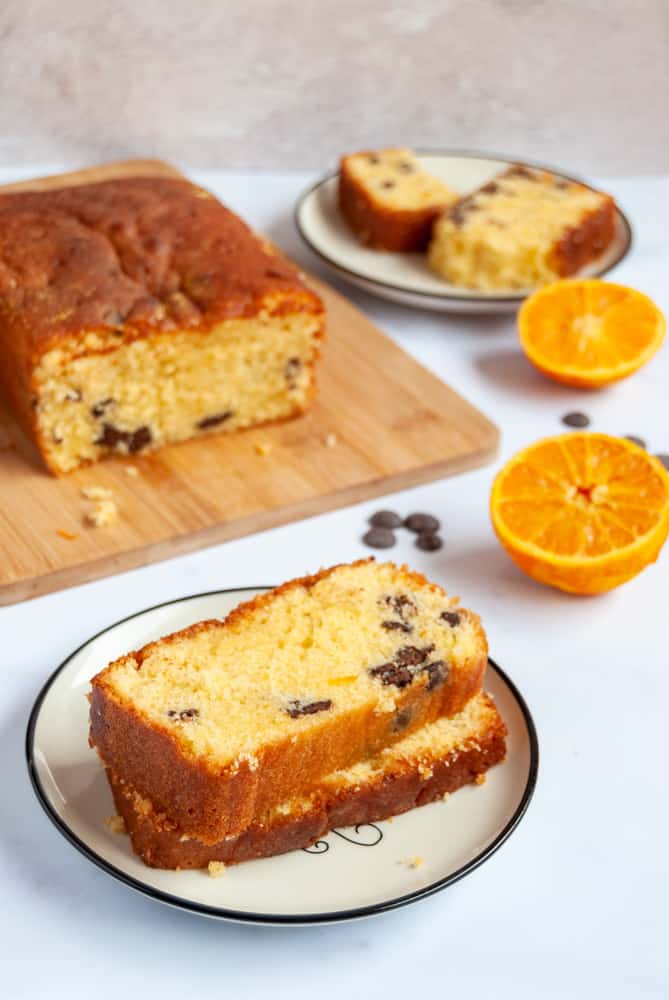 two slices of orange drizzle loaf cake with chocolate chips on a white and black plate. The rest of the cake is on a wooden board in the background along with an orange sliced in half and a plate with another slice of cake.