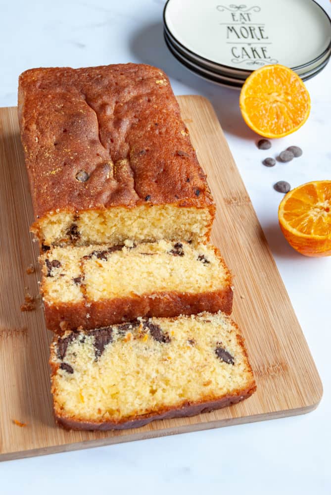 an orange loaf cake with chocolate chips on a wooden serving board. An orange cut in half and a sprinkling of chocolate chips are sitting beside the board along with a black and white serving plate. 