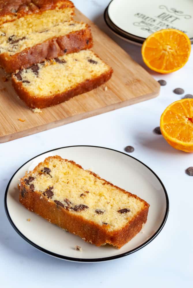 a slice of orange chocolate chip loaf cake on a white and black plate and an orange cake on a wooden chopping board.  An orange cut in half and a black and white plate are sitting beside the board.