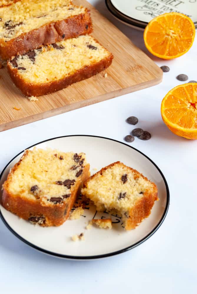 a slice of orange drizzle cake with chocolate chips on a black and white plate. A wooden board with an orange loaf cake and an orange sliced in half can be partially seen in the background.