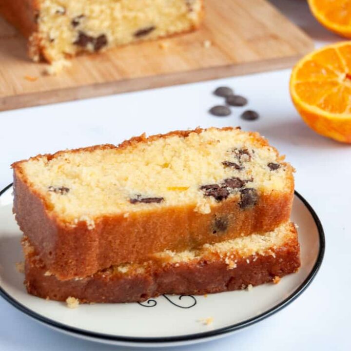 two slices of orange drizzle loaf cake on a white and black plate, an orange sliced in half, chocolate chips and an orange cake on a wooden board.