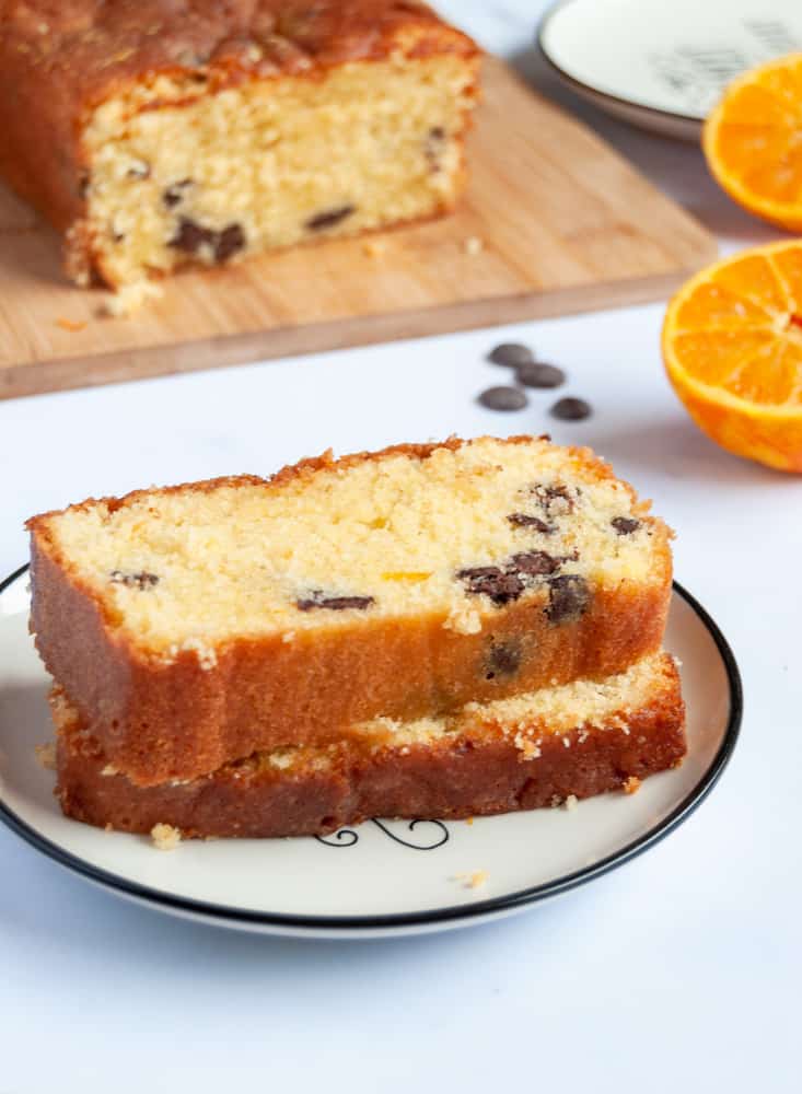 two slices of orange drizzle loaf cake on a white and black plate, an orange sliced in half, chocolate chips and an orange cake on a wooden board.