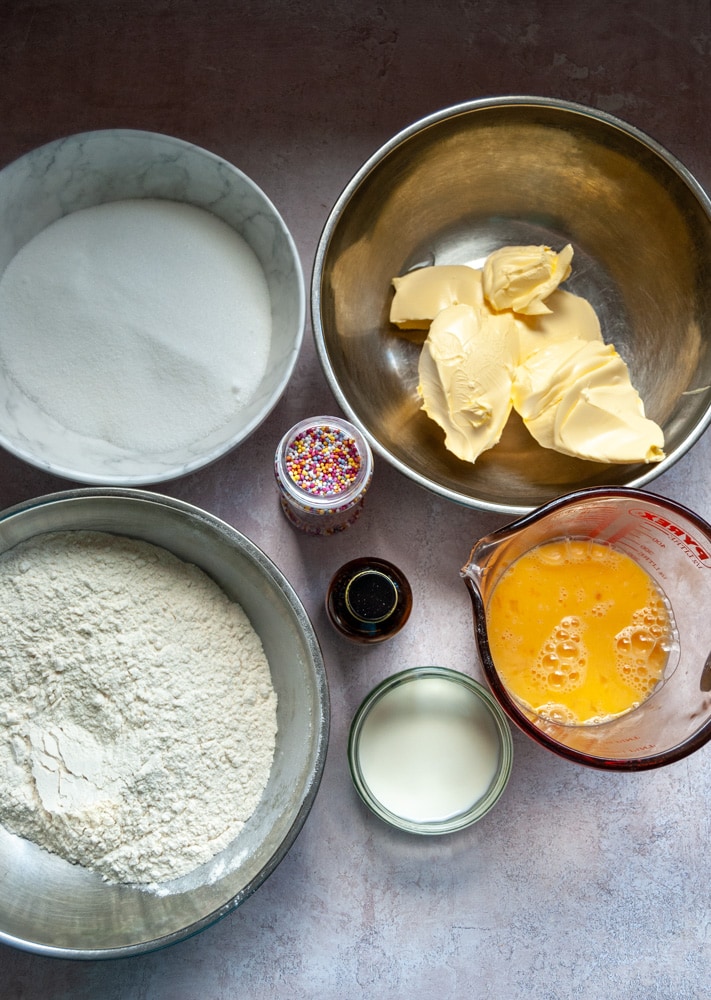 bowls of butter, sugar, flour, and a jug of cracked eggs, a small pot of milk and a bottle of vanilla extract and coloured sprinkles.