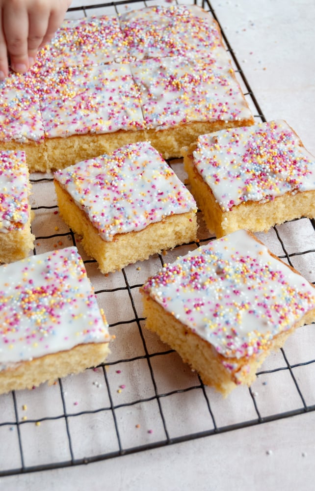 squares of vanilla sponge cake topped with white icing and coloured sprinkles on a black wire rack.  A toddler's hand decorating the cake with sprinkles can be partially seen in the top left corner .