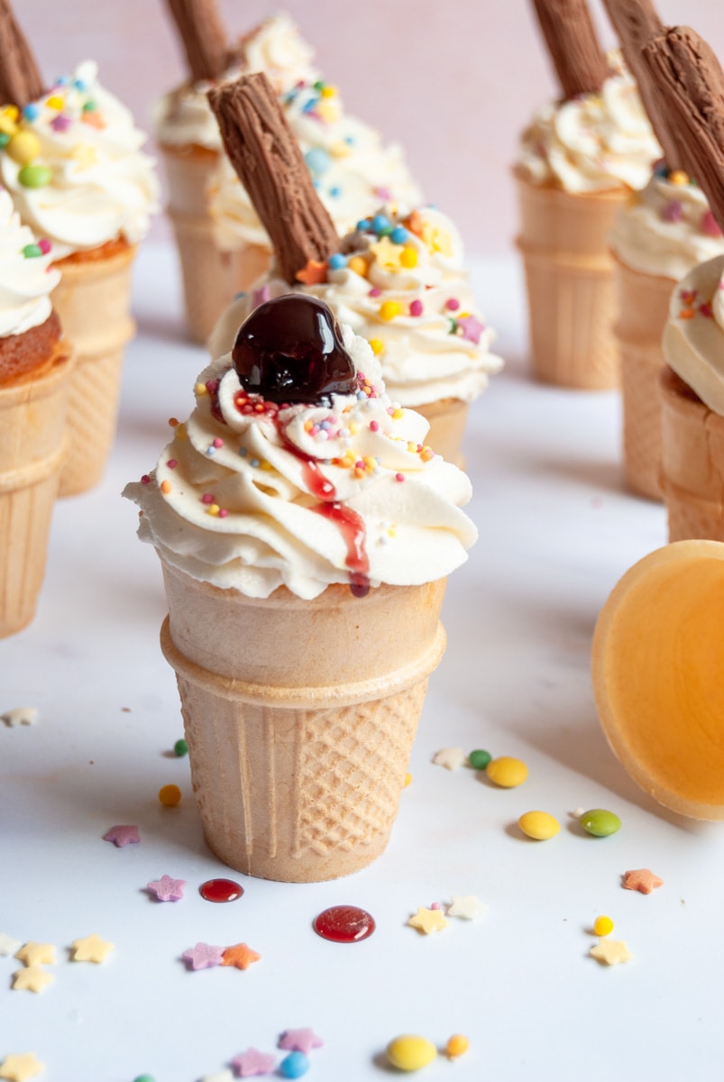 a flat bottomed ice cream cone cupcake topped with a vanilla buttercream swirl, coloured sprinkles and a marachino cherry. 