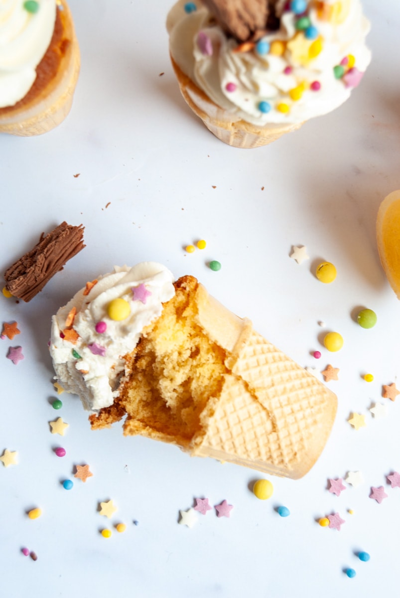 A vanilla cupcake baked into an ice cream cone lying on a white background, coloured sprinkles surrounding the cupcake.