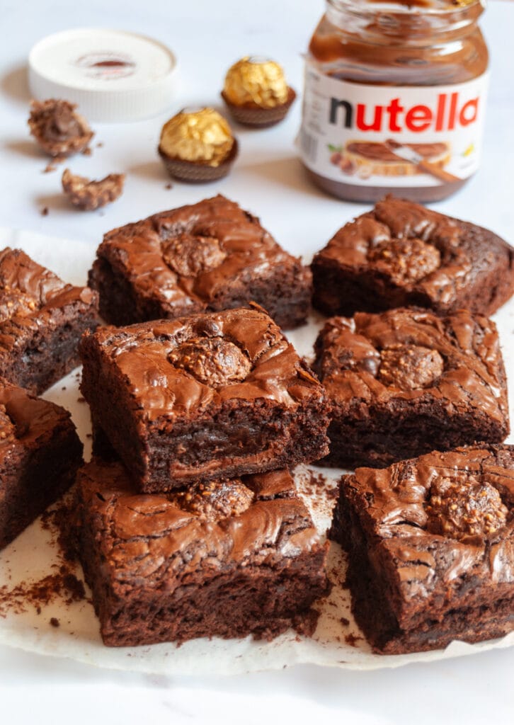 a batch of brownies filled with Nutella and topped with a ferrero rocher chocolate on a wooden board.  