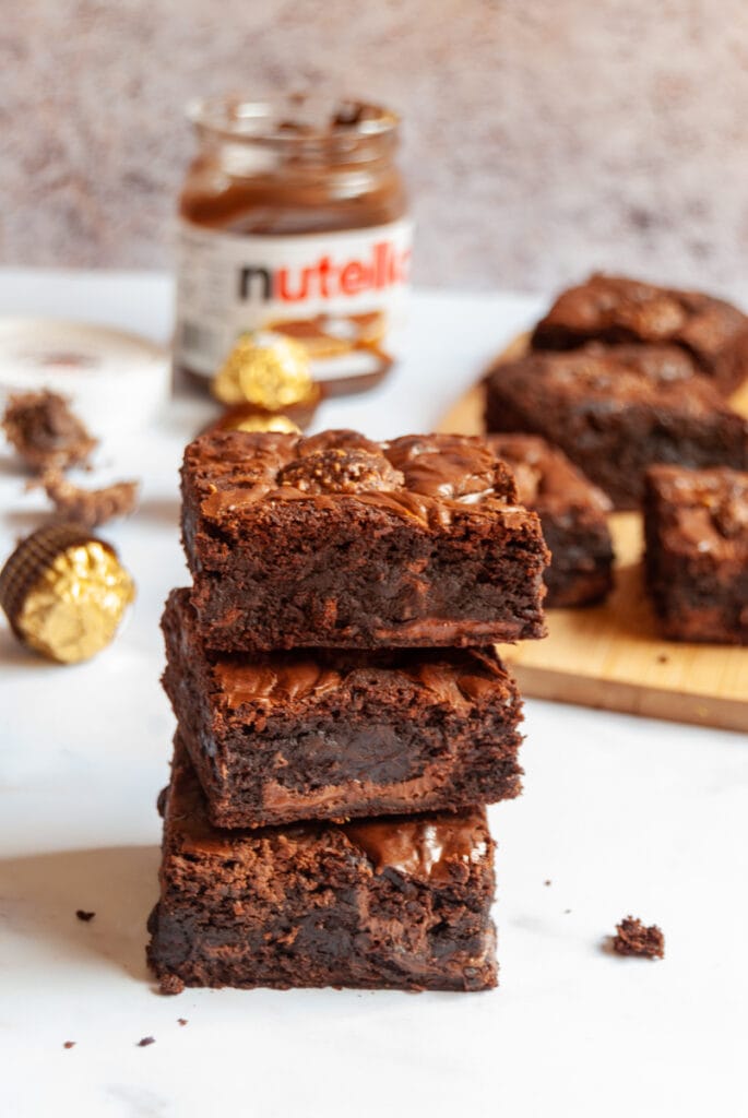 three brownies with Nutella swirls sitting on top of each other with a jar of Nutella in the background.