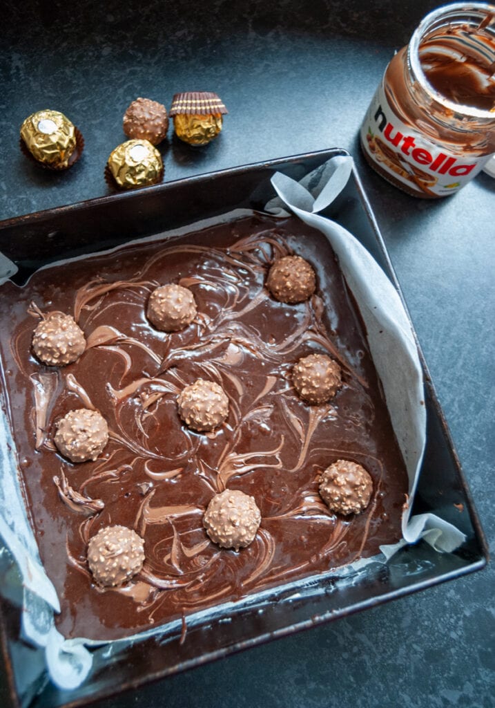 a square pan of unbaked brownies topped with ferrero rocher chocolates and a jar of nutella