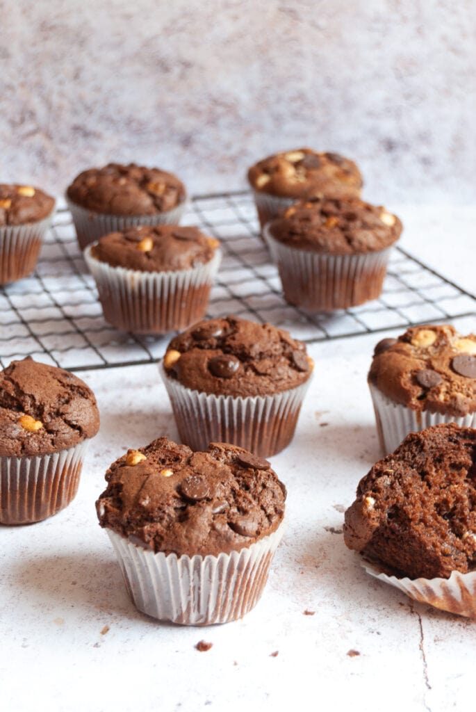 chocolate chip muffins on a white and grey backdrop. More muffins sit on a wire rack in the background.