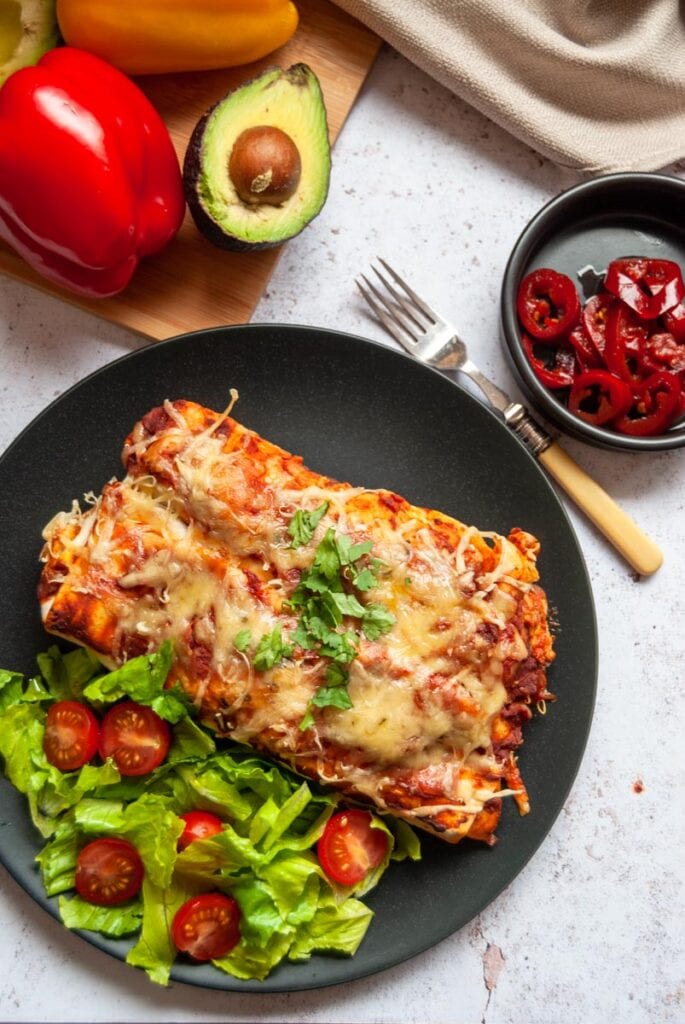 two chicken enchiladas with sliced lettuce and tomatoes on a black plate with a fork and a black bowl of red jalapeno peppers, an avocado and bell peppers.