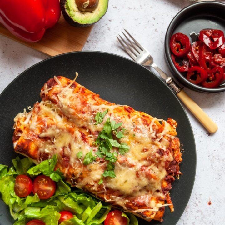 two chicken enchiladas with sliced lettuce and tomatoes on a black plate with a fork and a black bowl of red jalapeno peppers, an avocado and bell peppers.