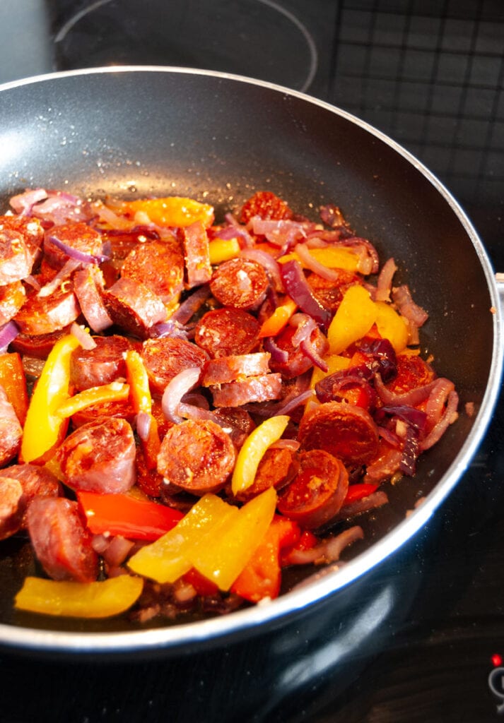 red onions, red and yellow sliced peppers and sliced chorizo frying in a pan.