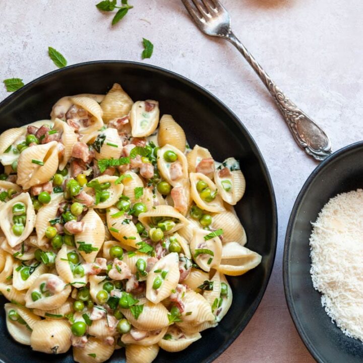 a bowl of shell pasta with peas, pancetta and fresh mint and a small bowl of parmesan cheese with a vintage style fork.