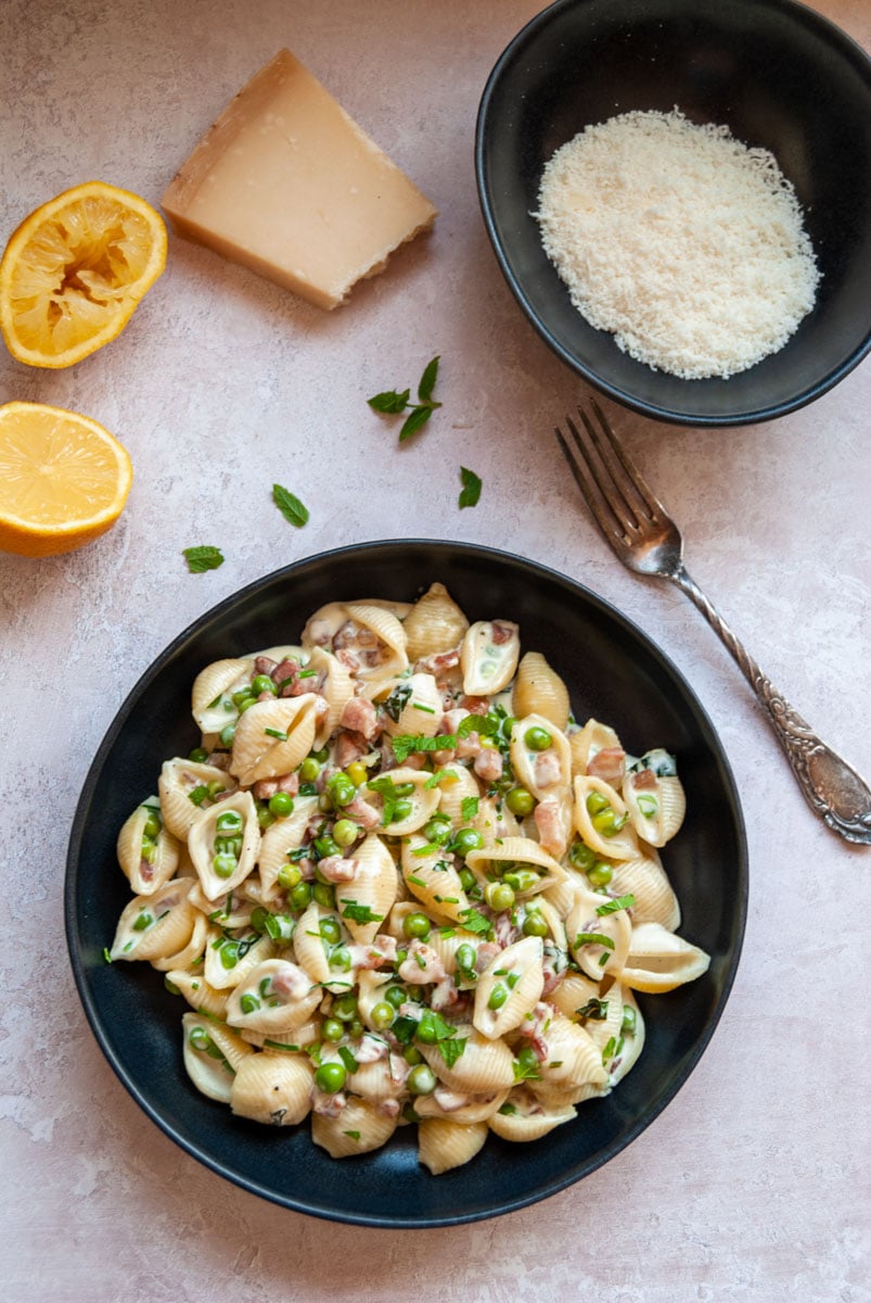 a bowl of pasta with pancetta, peas and cream, a silver fork, a bowl of grated parmesan and a lemon.