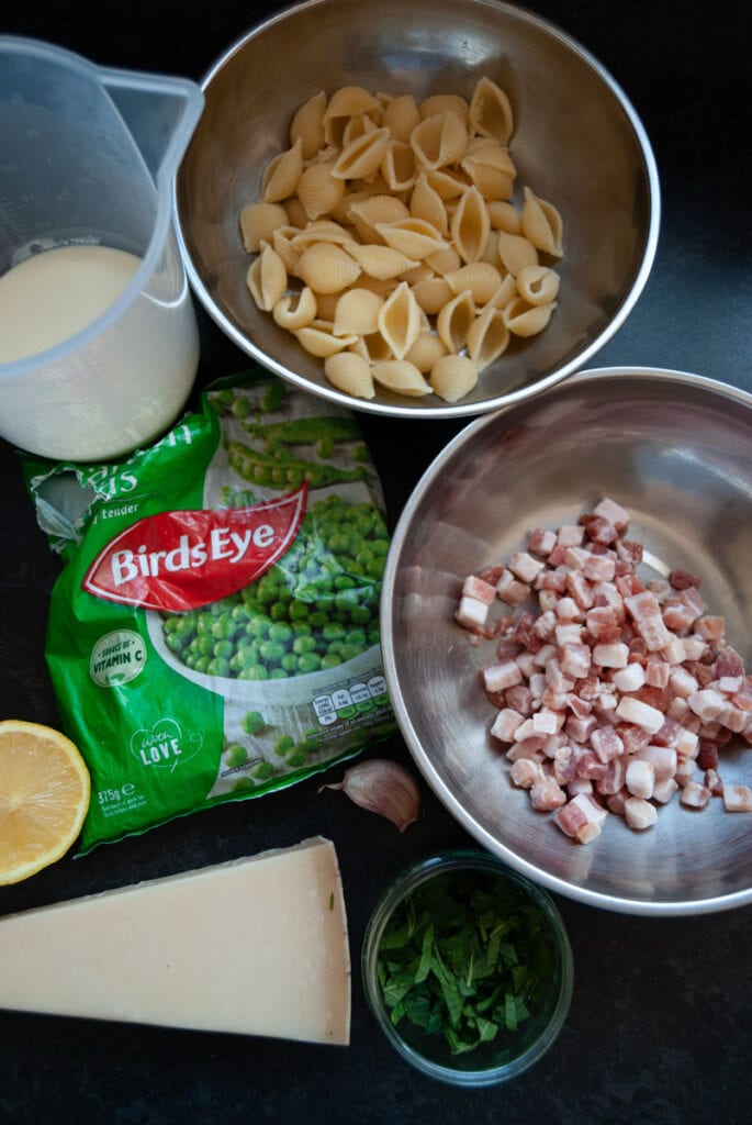 silver bowls of shell pasta, pancetta, a bag of frozen peas, a jug of cream, half a lemon, a bowl of fresh mint leaves and a wedge of parmesan cheese.