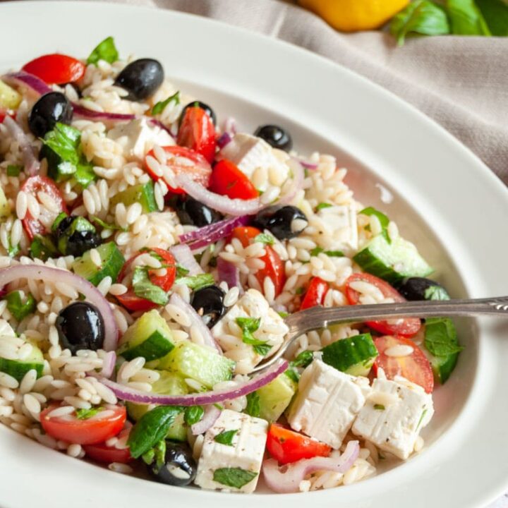 a greek salad with Orzo pasta on a white oval plate with a serving spoon.