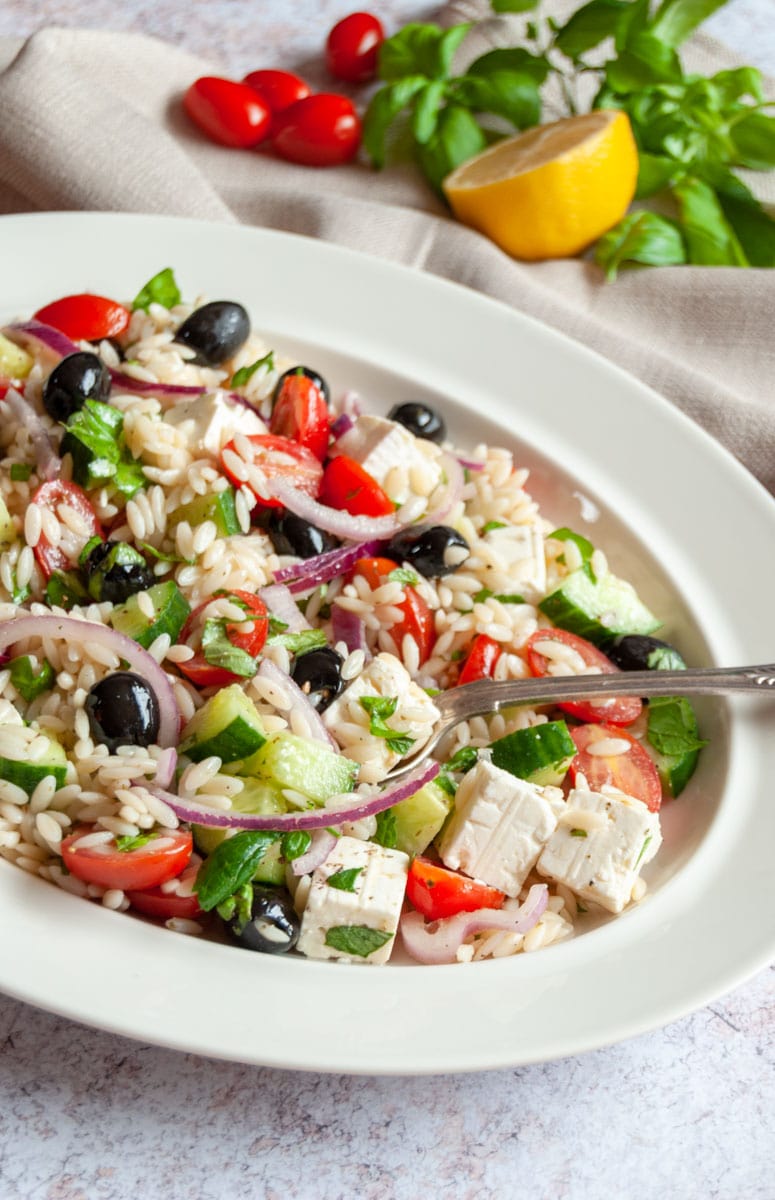 a greek salad with Orzo pasta on a white oval plate with a serving spoon.