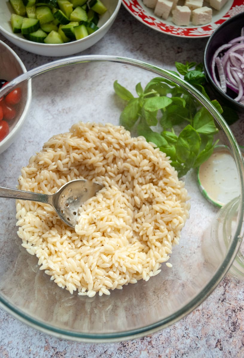 A glass bowl of orzo pasta with a spoon, chopped herbs and bowls of red onion, feta cheese, chopped cucumber and cherry tomatoes.