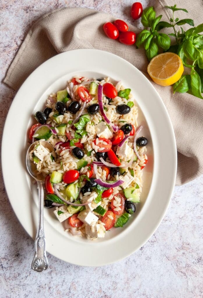an orzo salad with black olives, cherry tomatoes, red onions and herbs on a white oval plate with a serving spoon.