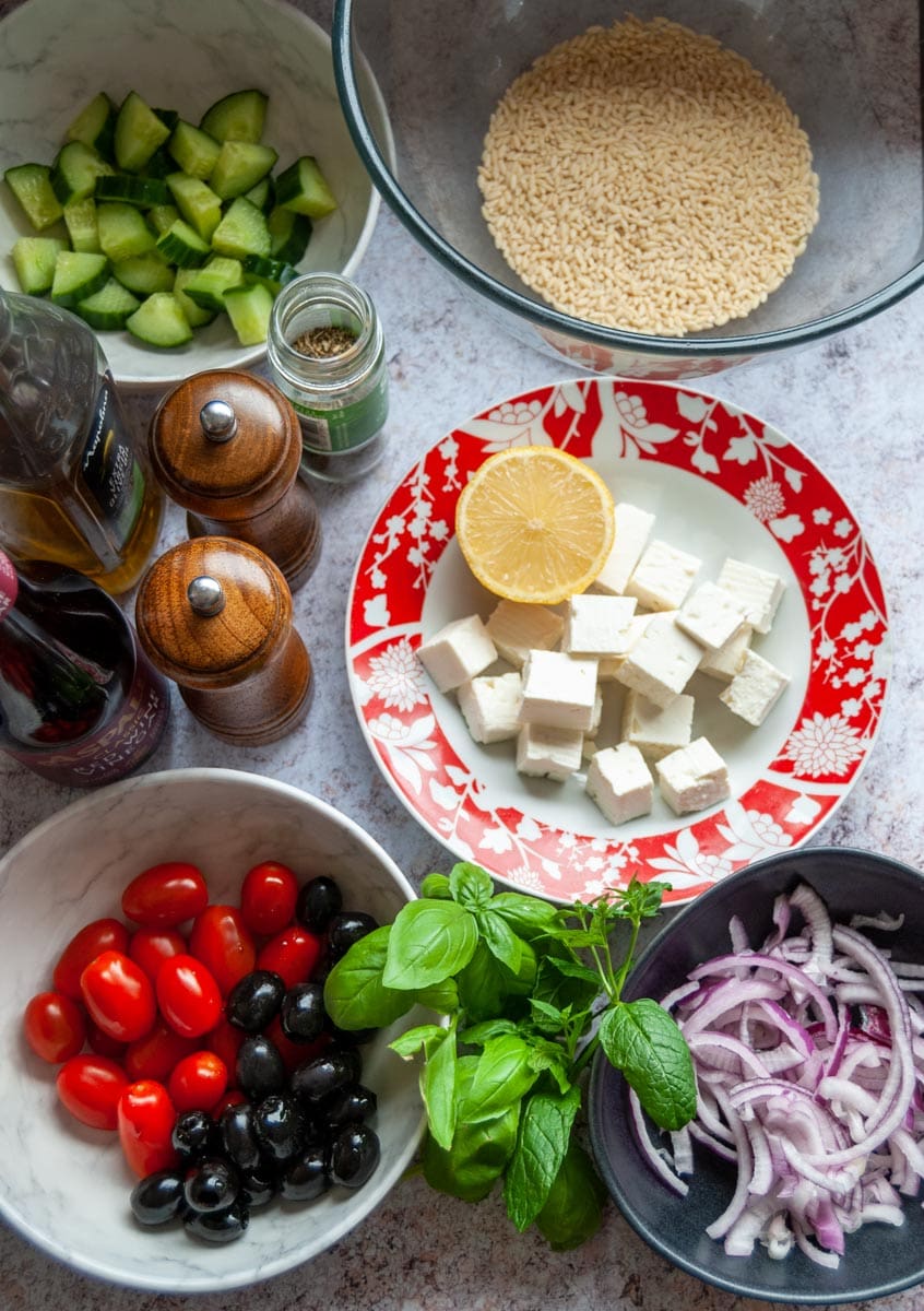 bowls of cherry tomatoes, black olives, sliced red onion, feta cheese, fresh herbs, chopped cucumber, orzo pasta and a wooden salt and pepper mill.