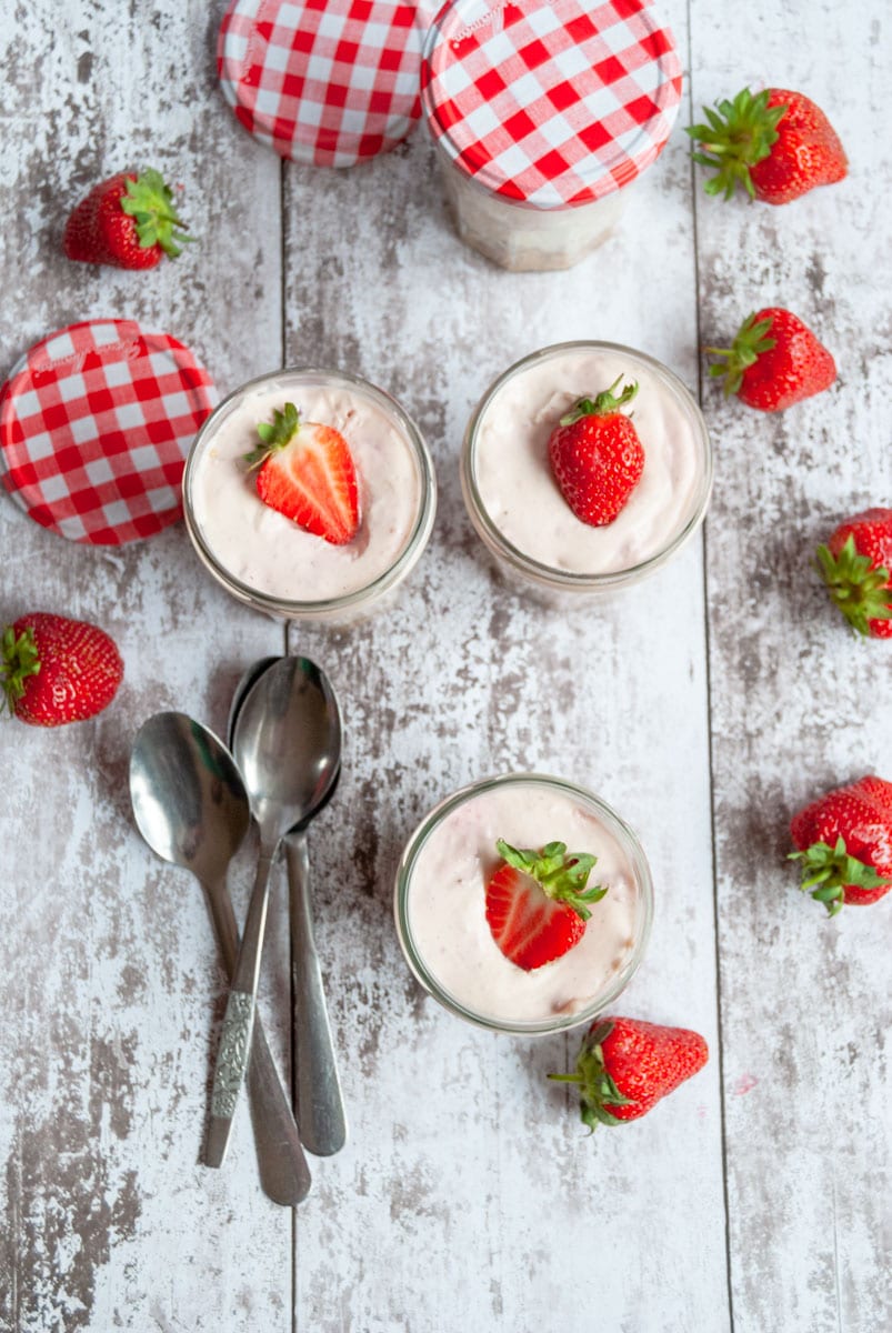 three strawberry cheesecake in a jar topped with a fresh sliced strawberry with silver spoons on a distressed grey and white wooden board.