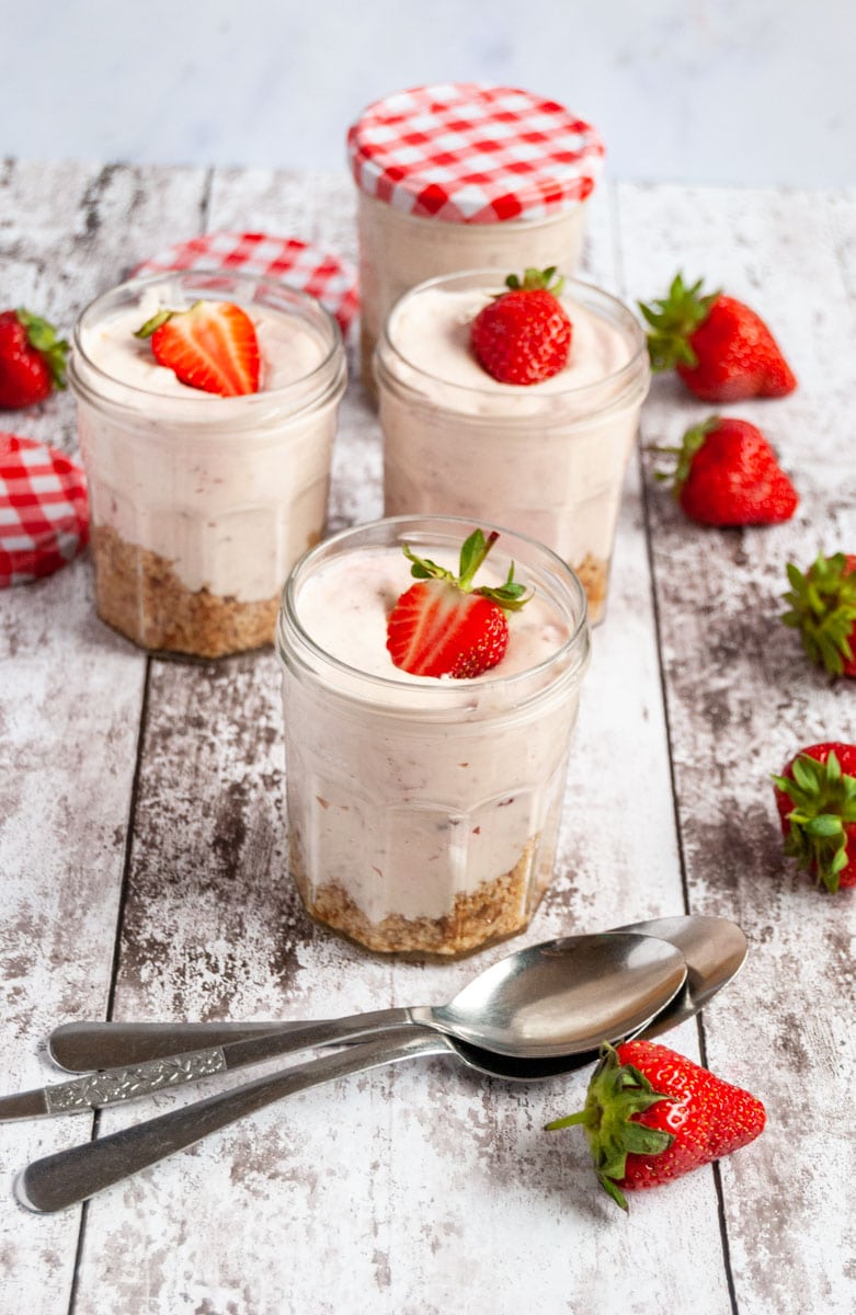 three jars of strawberry cheesecake topped with a fresh sliced strawberry, silver spoons and fresh strawberries on a distressed white and grey wooden board.