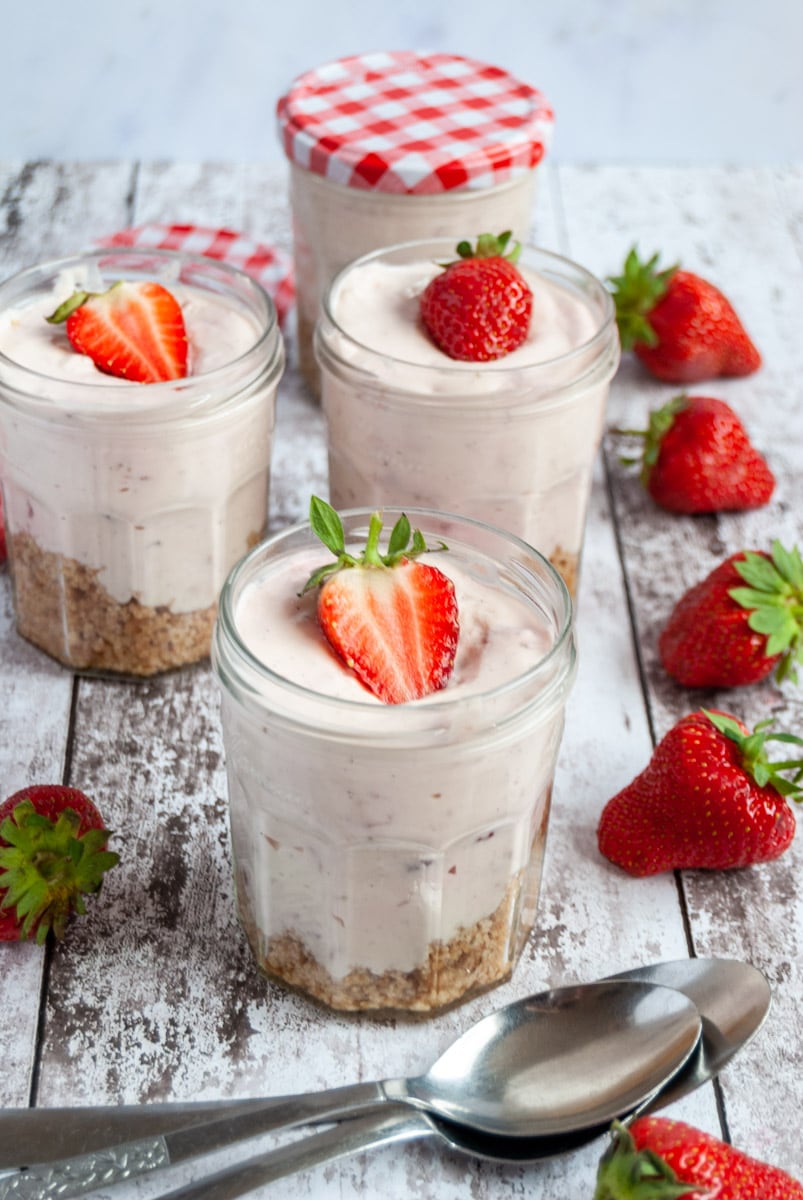 three no bake strawberry cheesecakes in jam jars topped with a fresh strawberry and silver spoons on a white and grey wooden board.