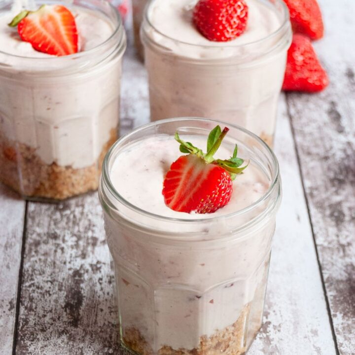 three no bake strawberry cheesecakes in jam jars topped with a fresh strawberry