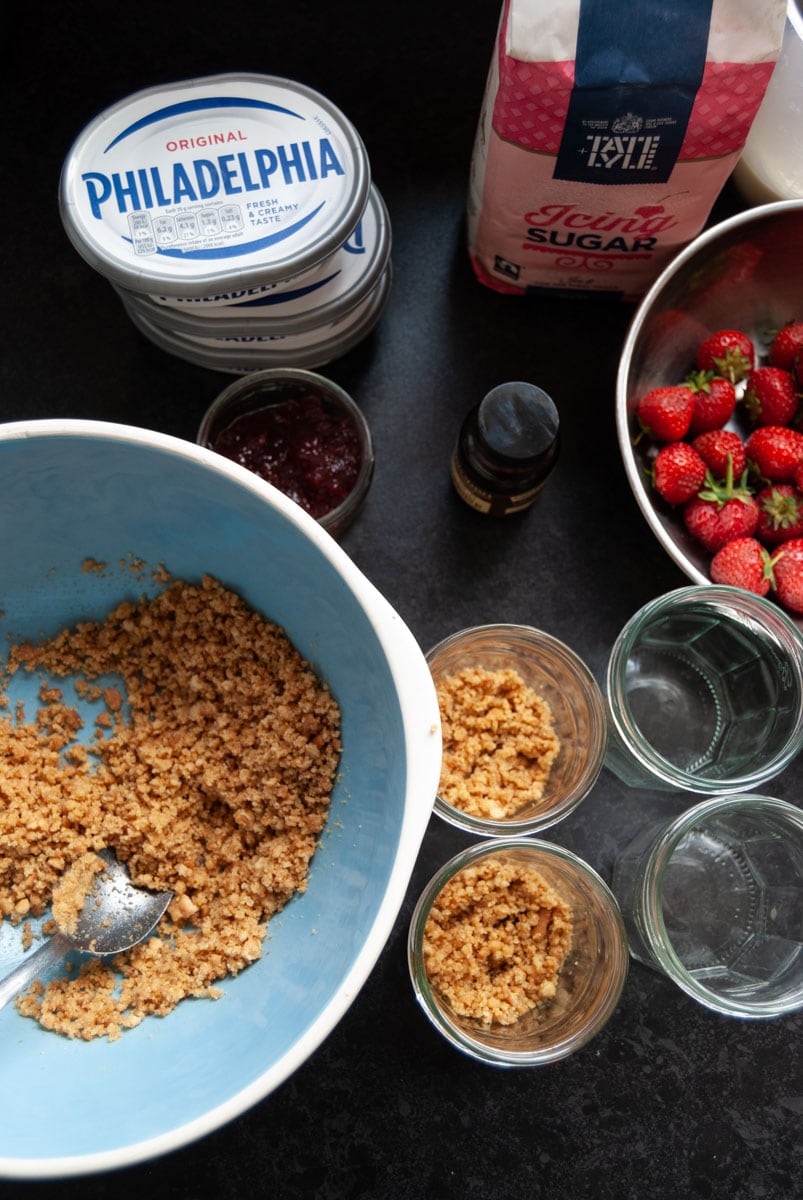 tubs of cream cheese, a blue and white bowl with crushed biscuits, a bowl of strawberries, a bag of icing sugar and jam jars filled with crushed biscuits.