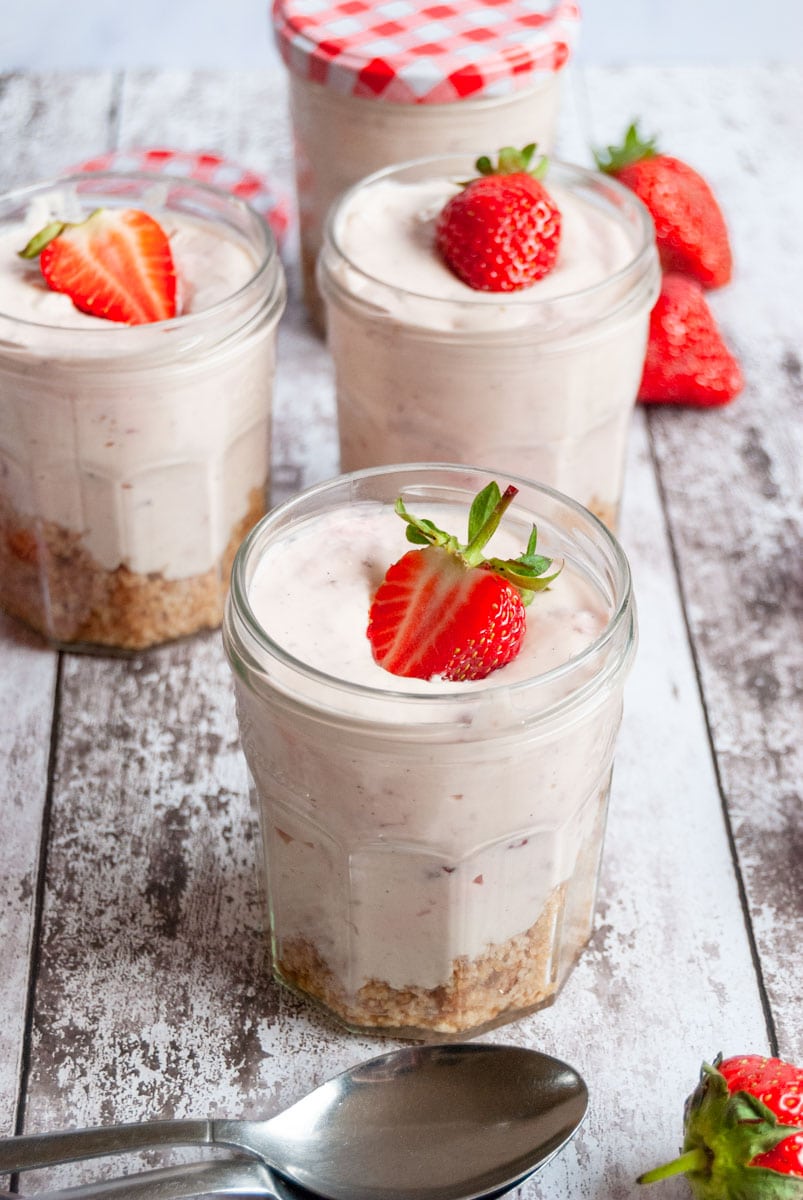 three no bake strawberry cheesecakes in jam jars topped with a fresh strawberry