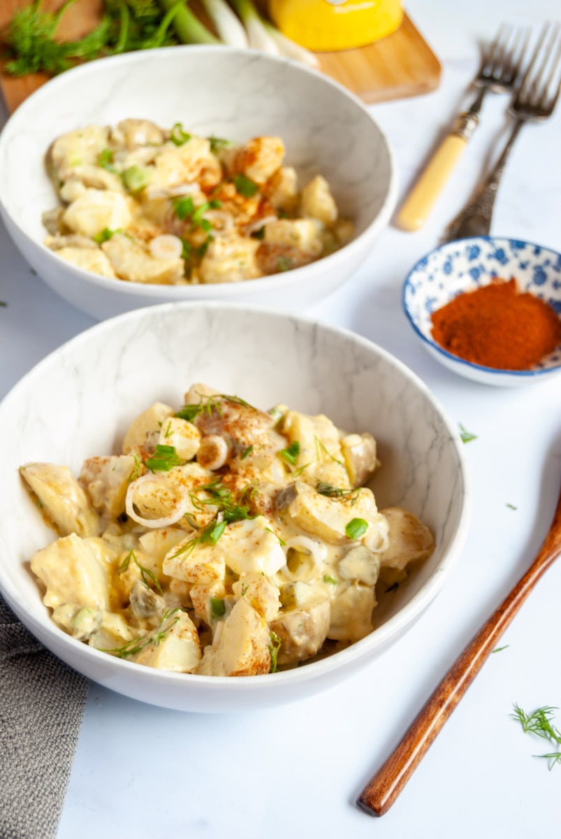 two white bowls of potato and egg salad with sliced sping onions and fresh dill, a wooden spoon, two forks and a blue pot of paprika.