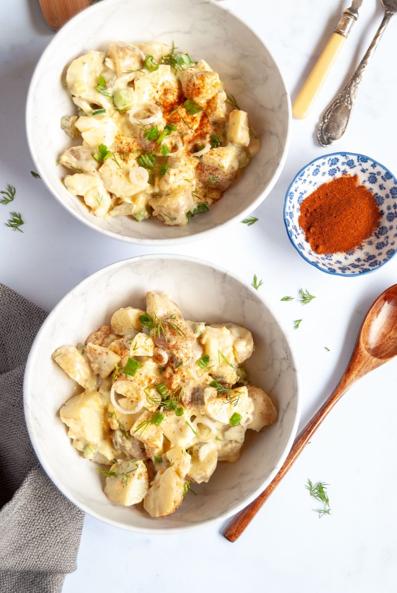 two white bowls of potato salad with chopped eggs, spring onions and freshly chopped dill, two forks, a wooden spoon and a small blue pot of paprika.