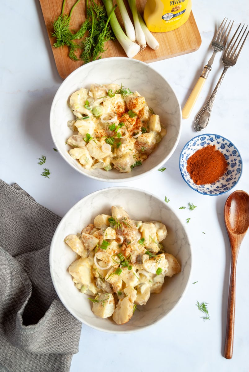 two white bowls of potato salad with chopped boiled eggs, Spring onions and fresh dill, a brown wooden spoon and a blue dish of paprika with two forks. 