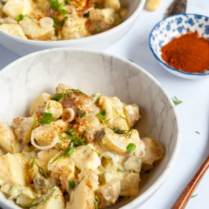 two small white bowls of potato salad with chopped boiled eggs, sliced spring onions and fresh dill and small blue dish of paprika