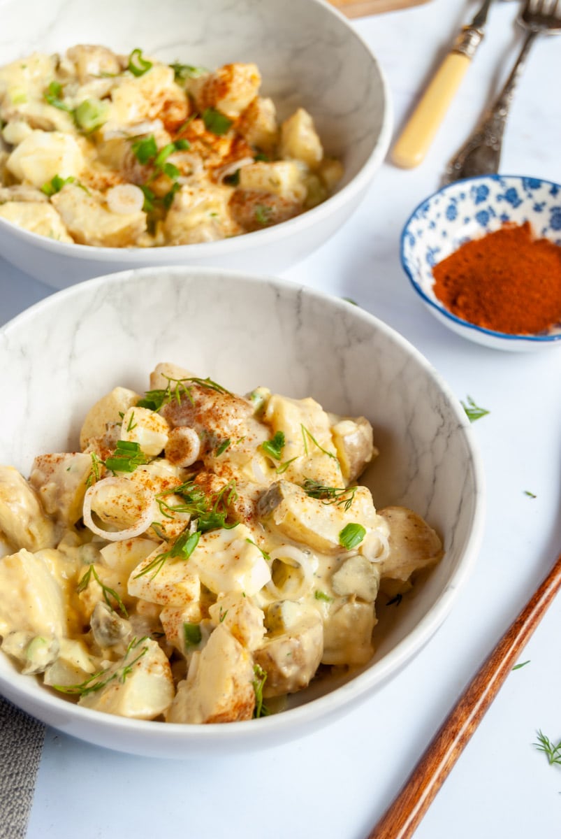 two small white bowls of potato salad with chopped boiled eggs, sliced spring onions and fresh dill and small blue dish of paprika