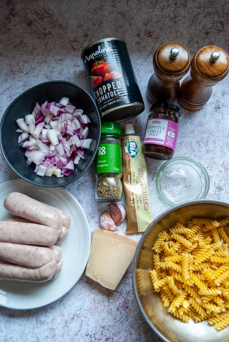 a silver bowl of pasta, a white plate of sausages, a bowl of sliced red onion, a can of chopped tomatoes, parmesan cheese, a jar of chilli paste, a jar of dried oregano and salt and pepper mills.