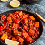 a black bowl of sweet potato chilli with red peppers and black beans, lime wedges and a small bowl of Mexican rice.