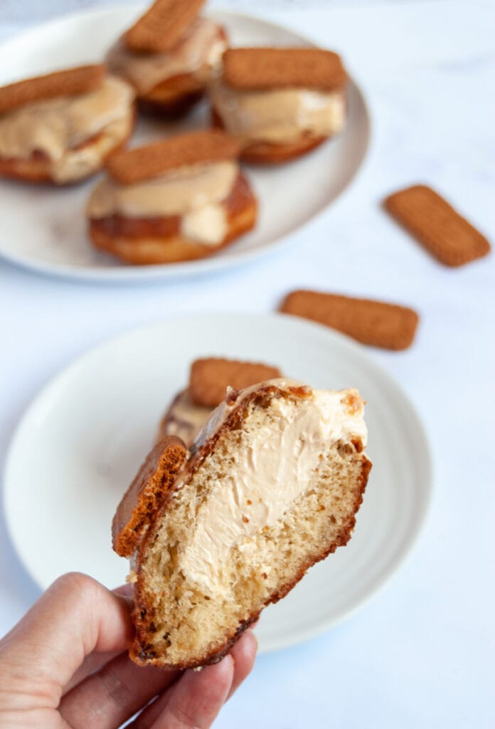 a hand holding half a doughnut filled with biscoff whipped cream and topped with Biscoff glaze and a Biscoff cookie.  More doughnuts on a white plate can be seen in the background.