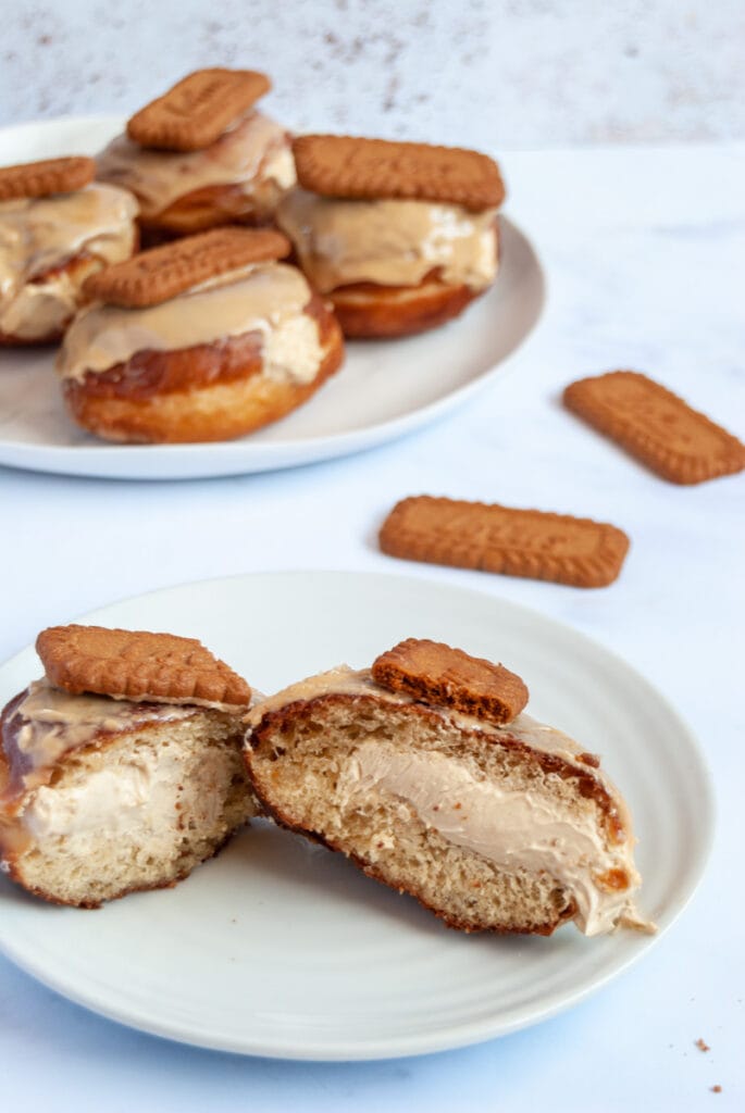 A doughnut filled with fresh cream and topped with a Biscoff biscuit cut in half on a white plate.  More doughnuts can be seen on a larger white plate in the background.