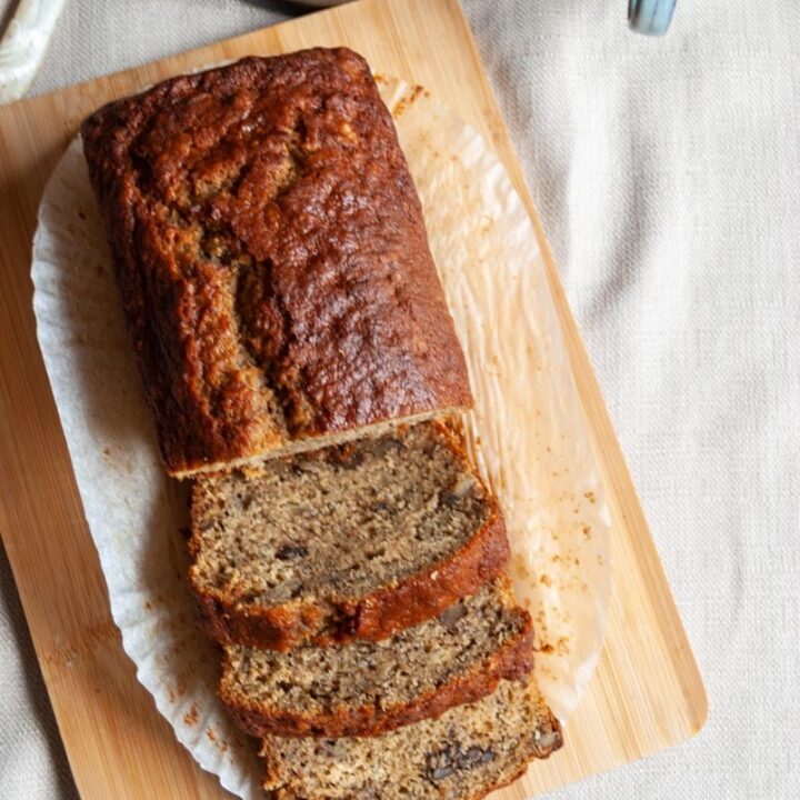 a banana loaf cake on a wooden board, a pot of butter and a light blue cup of coffee on a beige linen napkin.